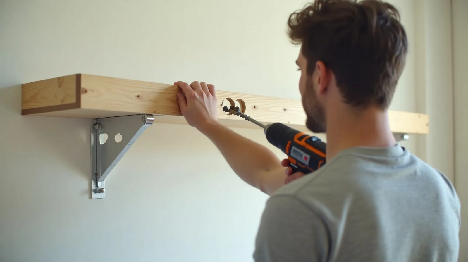 A person seen from behind driving a screw through a shelf bracket into a marked wall with a cordless drill, a spirit level tucked under one arm